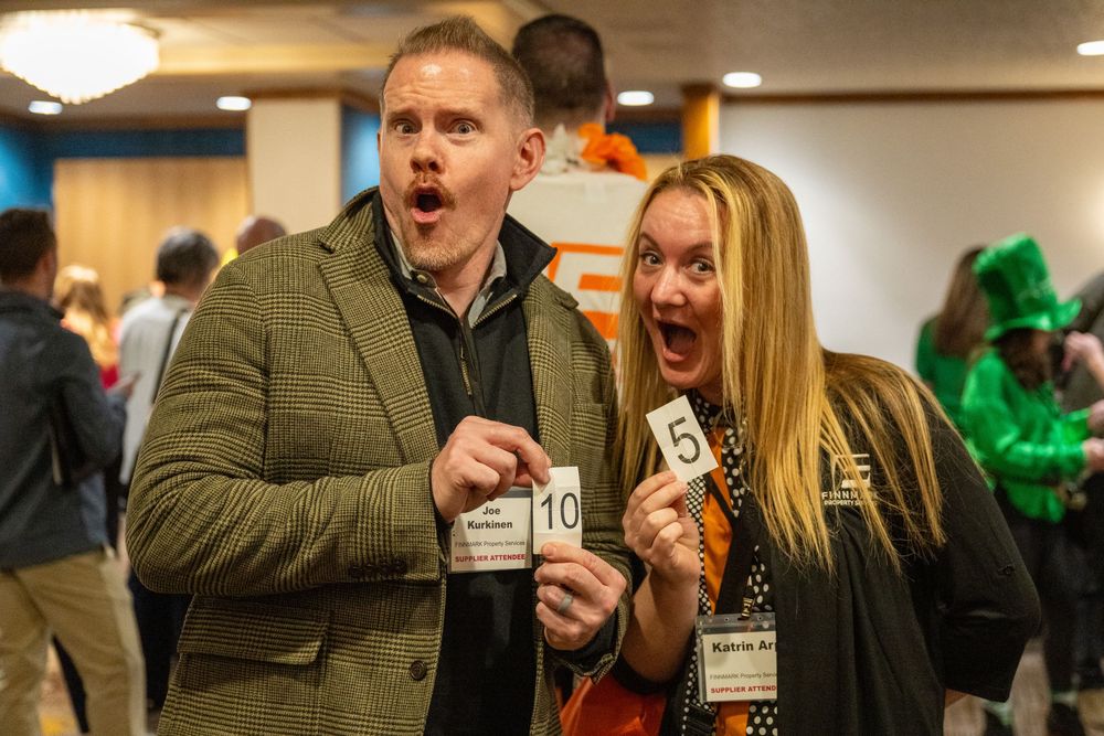 Two excited attendees showing off their tickets at an event with festive green decorations in the background.