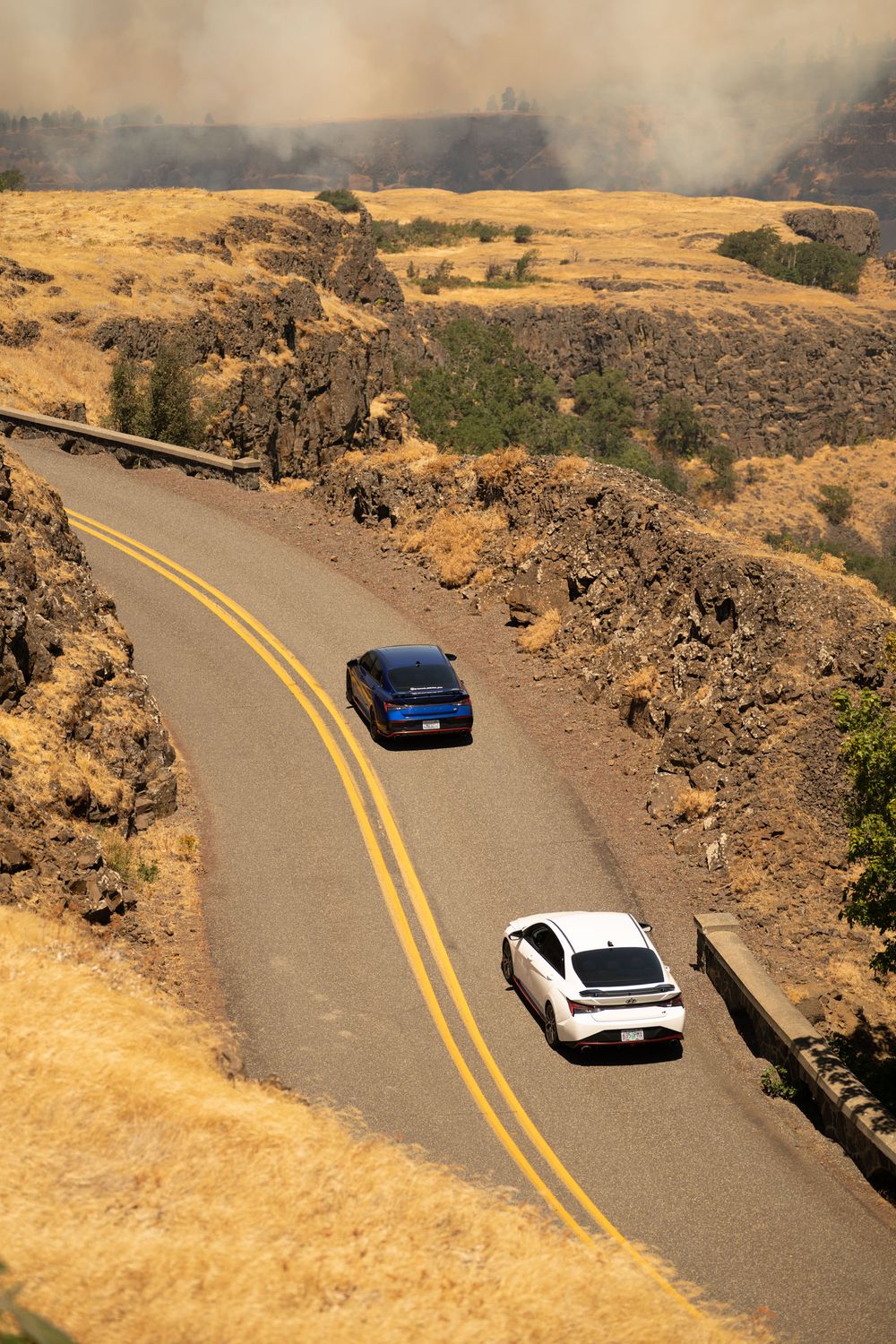 Cars drive along a winding mountain road with golden hills and rock formations in the background.