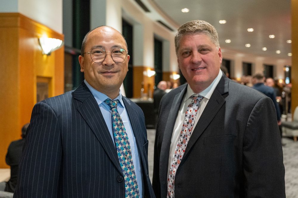 Two people in formal business attire stand together at a professional event in an indoor venue.