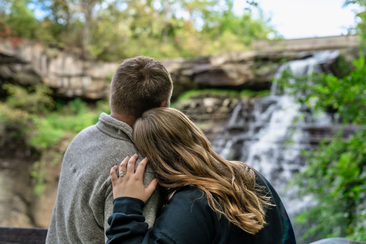 A couple shares a tender moment while sitting on rocks in front of scenic Brandywine Falls in Ohio for a surprise engagement photo session.