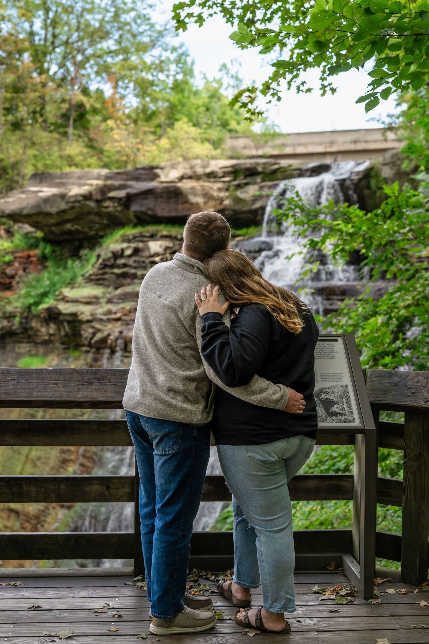 Couple embracing on nature boardwalk with scenic Brandywine Falls in Ohio for surprise engagement photo session.