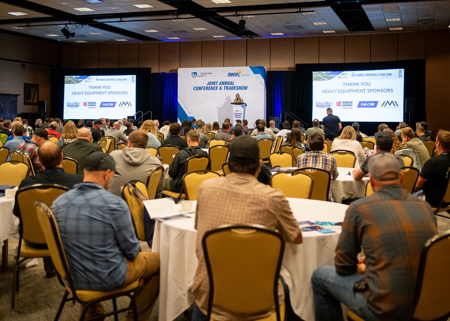 Conference attendees seated at round tables facing a stage with presentation screens.