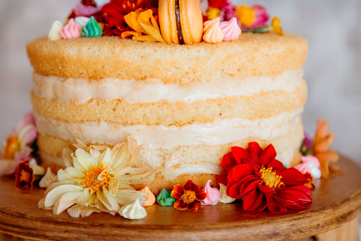 A naked rustic layer cake decorated with colorful fresh flowers and macarons on top sits on a wooden board.