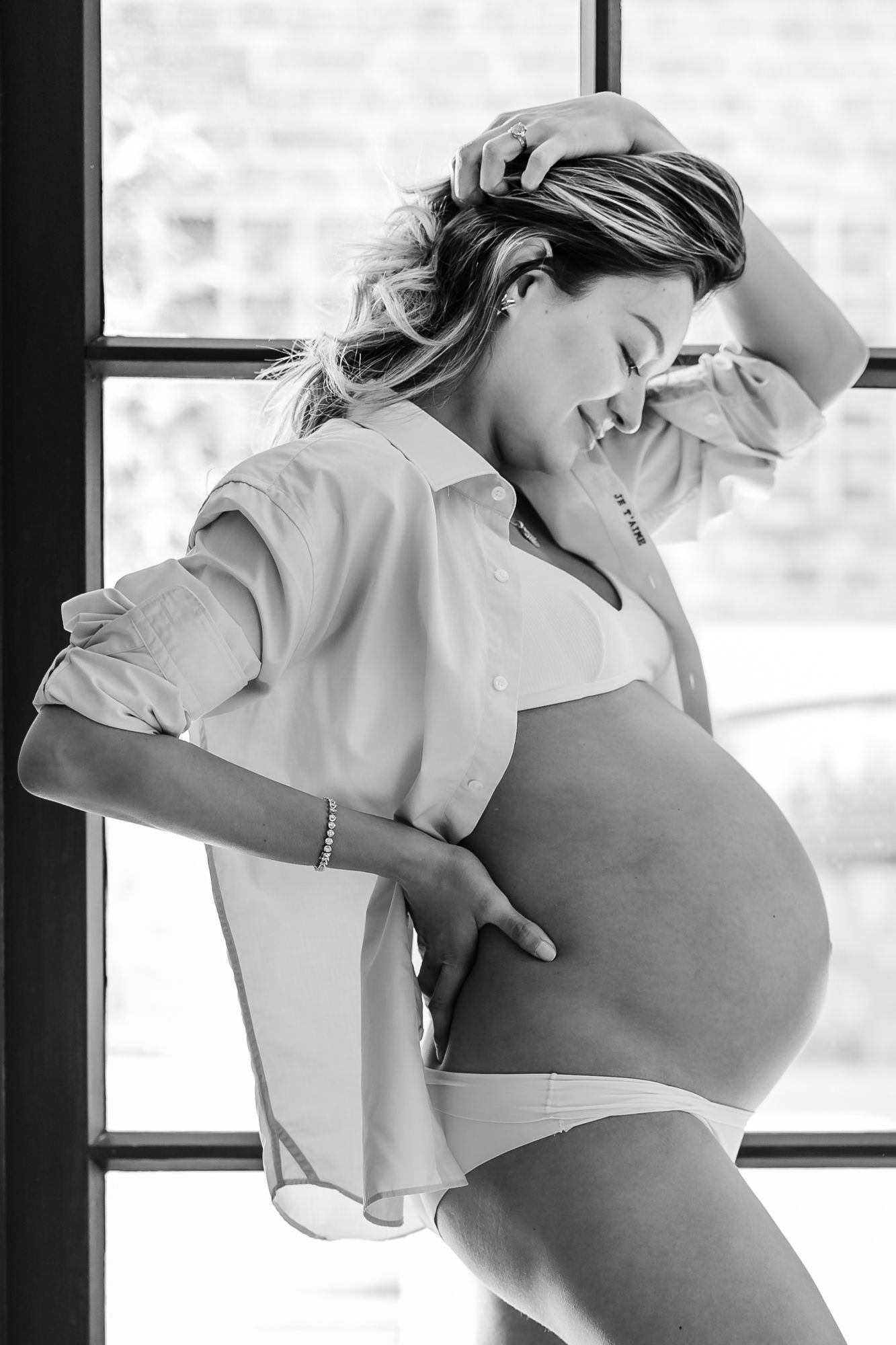 A black and white maternity photo shows an expectant mother in a white shirt by a window, gently touching her belly.