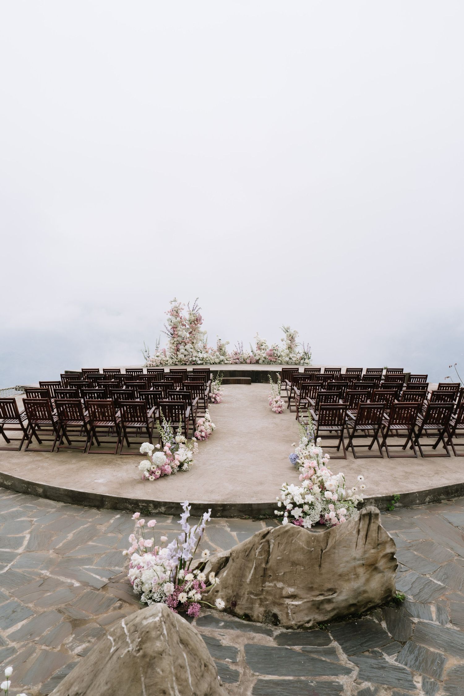 Elegant outdoor wedding ceremony venue on a stone terrace with floral aisle decor and misty sky.