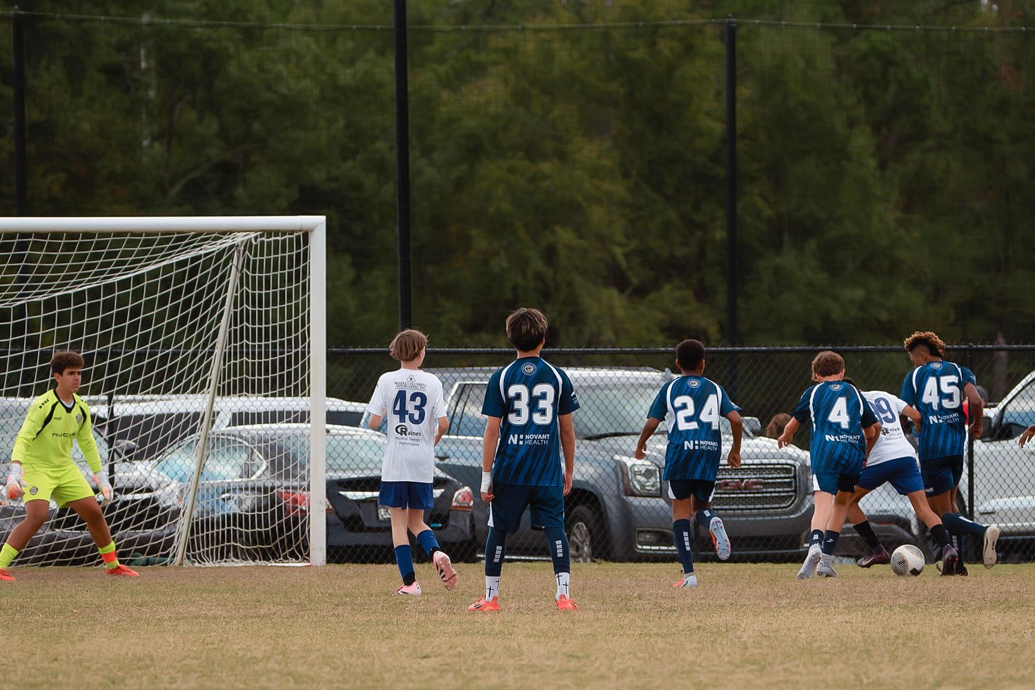 Soccer players sit on the ground in a row watching a game from the sidelines with a goal visible in the background.