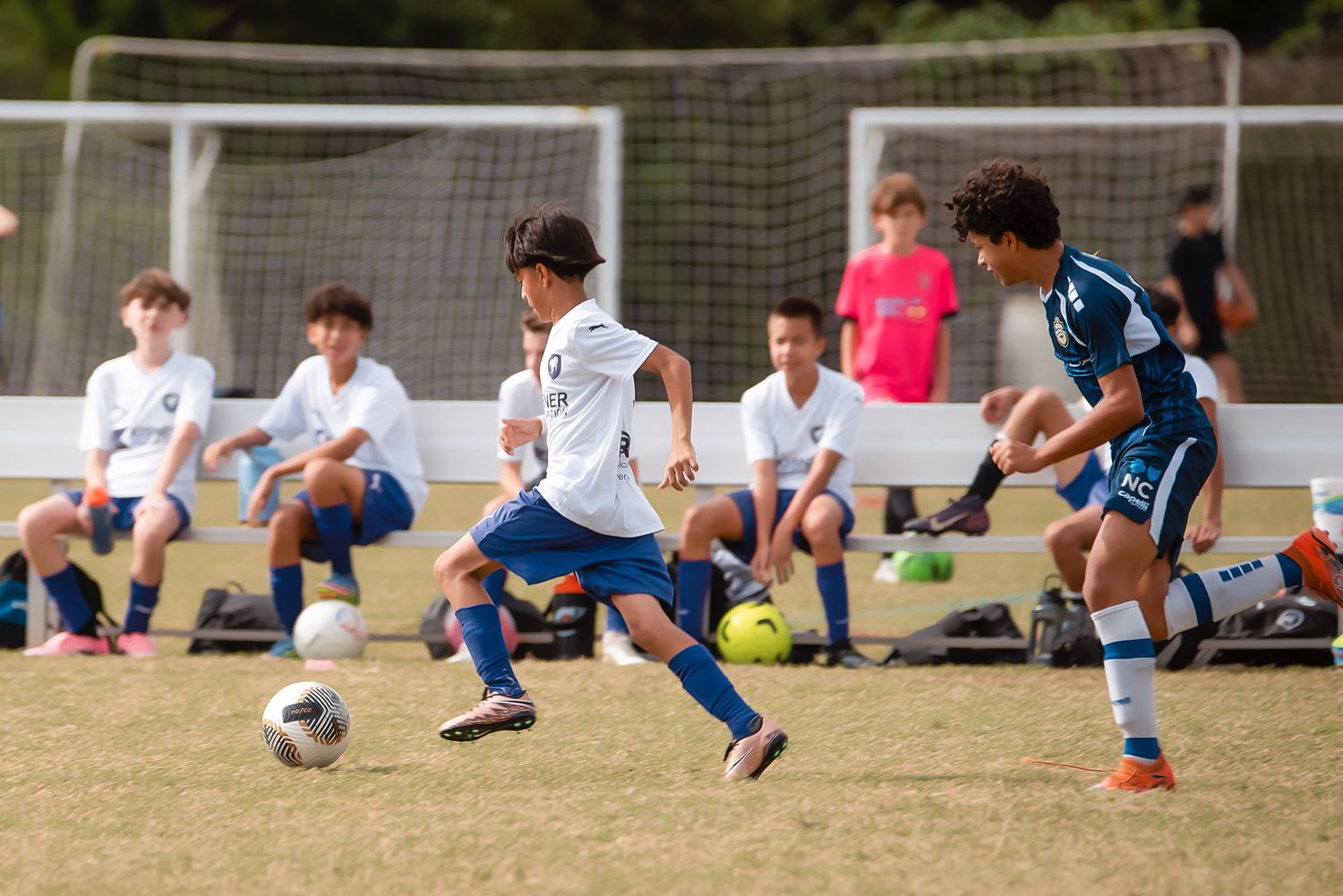 A row of soccer players practicing dribbling drills on a sunny grass field during training.