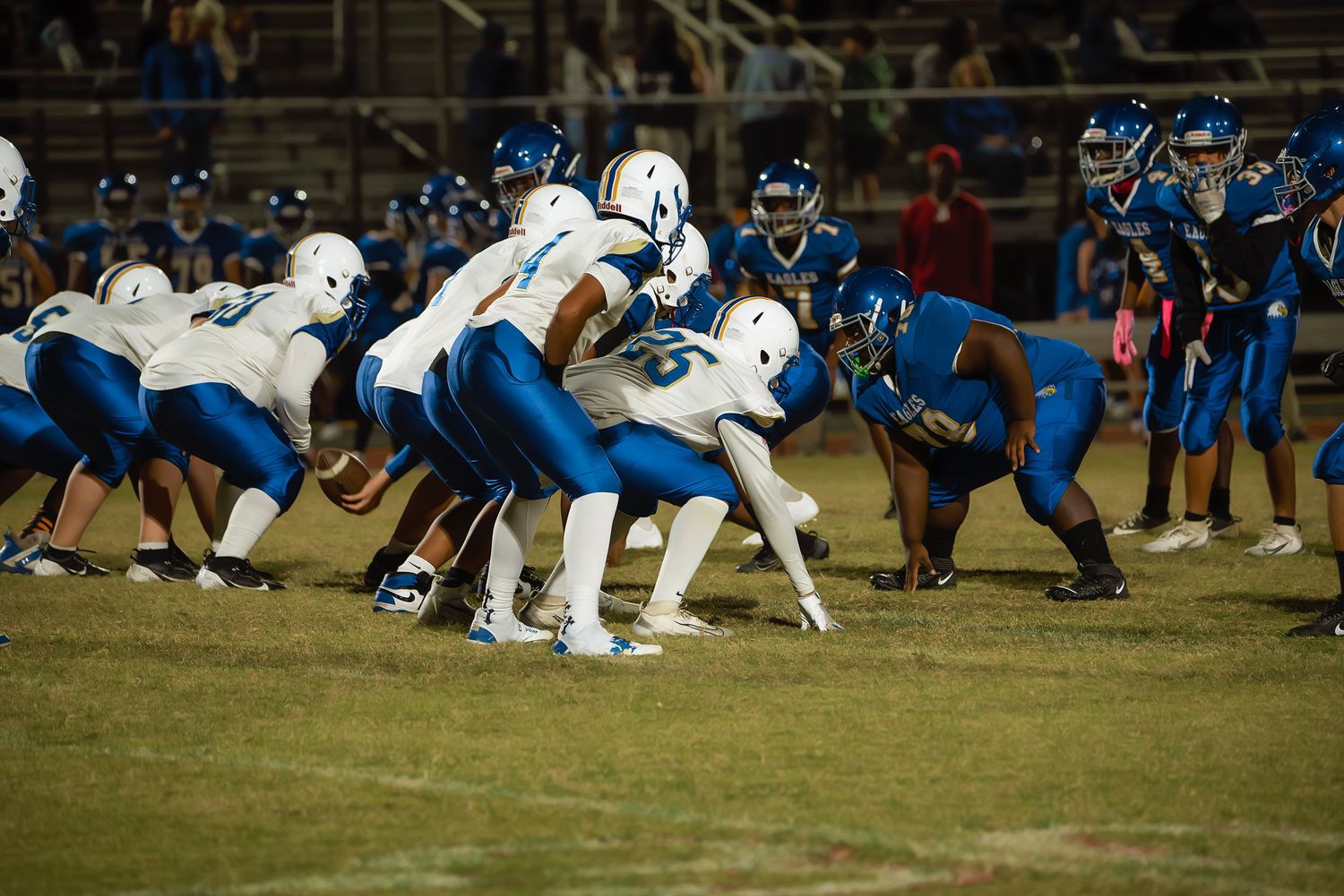 Football players from opposing teams in blue and white uniforms line up at scrimmage during a night game.