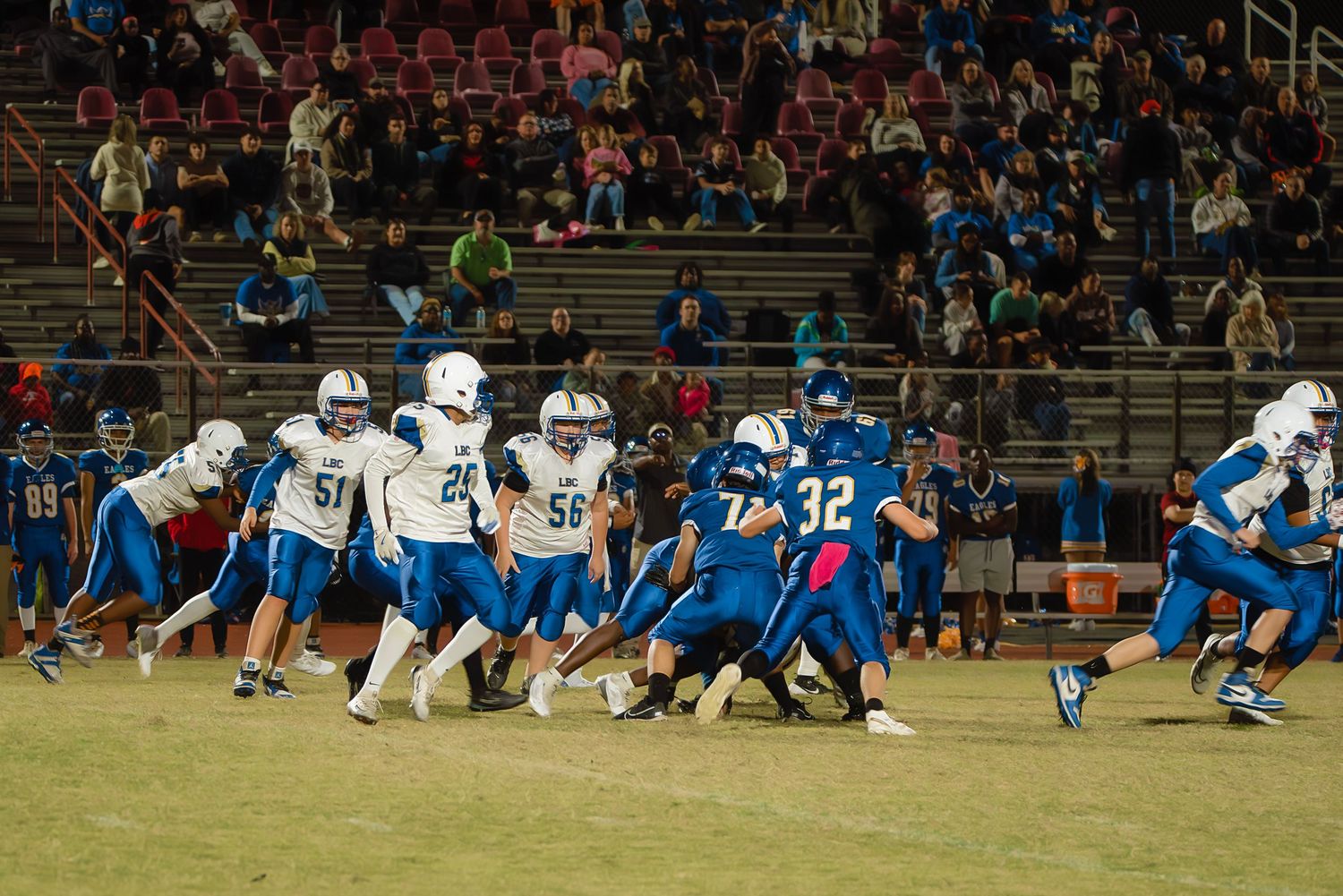 High school football teams in white and blue uniforms face off during a nighttime game on the field.