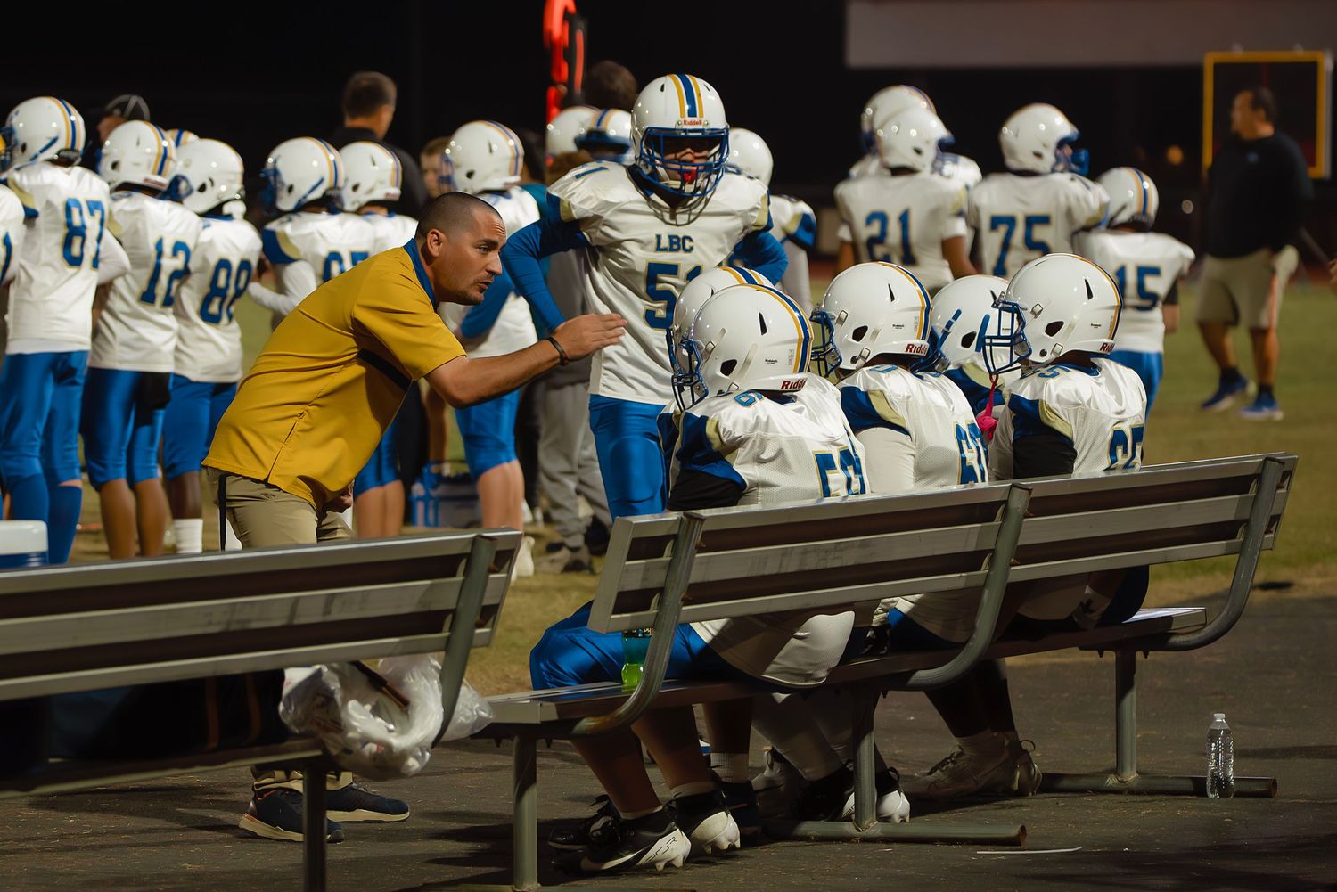 Row of football players in white uniforms sit on the sideline bench during a game.