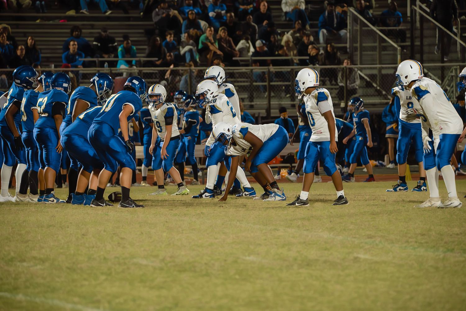 High school football players in blue uniforms line up for a play during a night game under stadium lights.