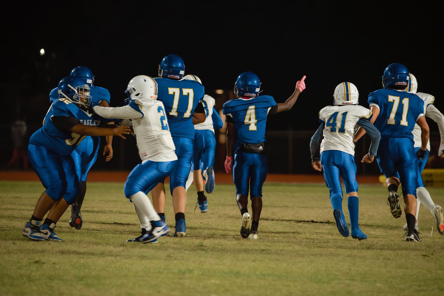 Football players in blue and white uniforms compete during a night game on an illuminated field.