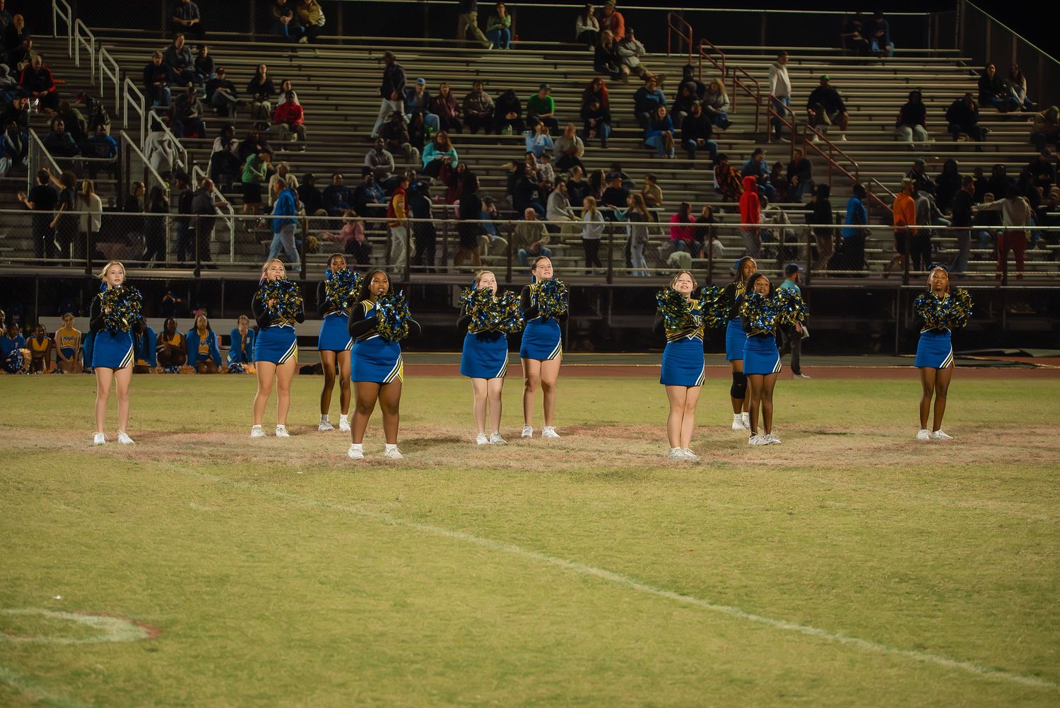Football players in blue uniforms line up on a field during a nighttime game under stadium lights.