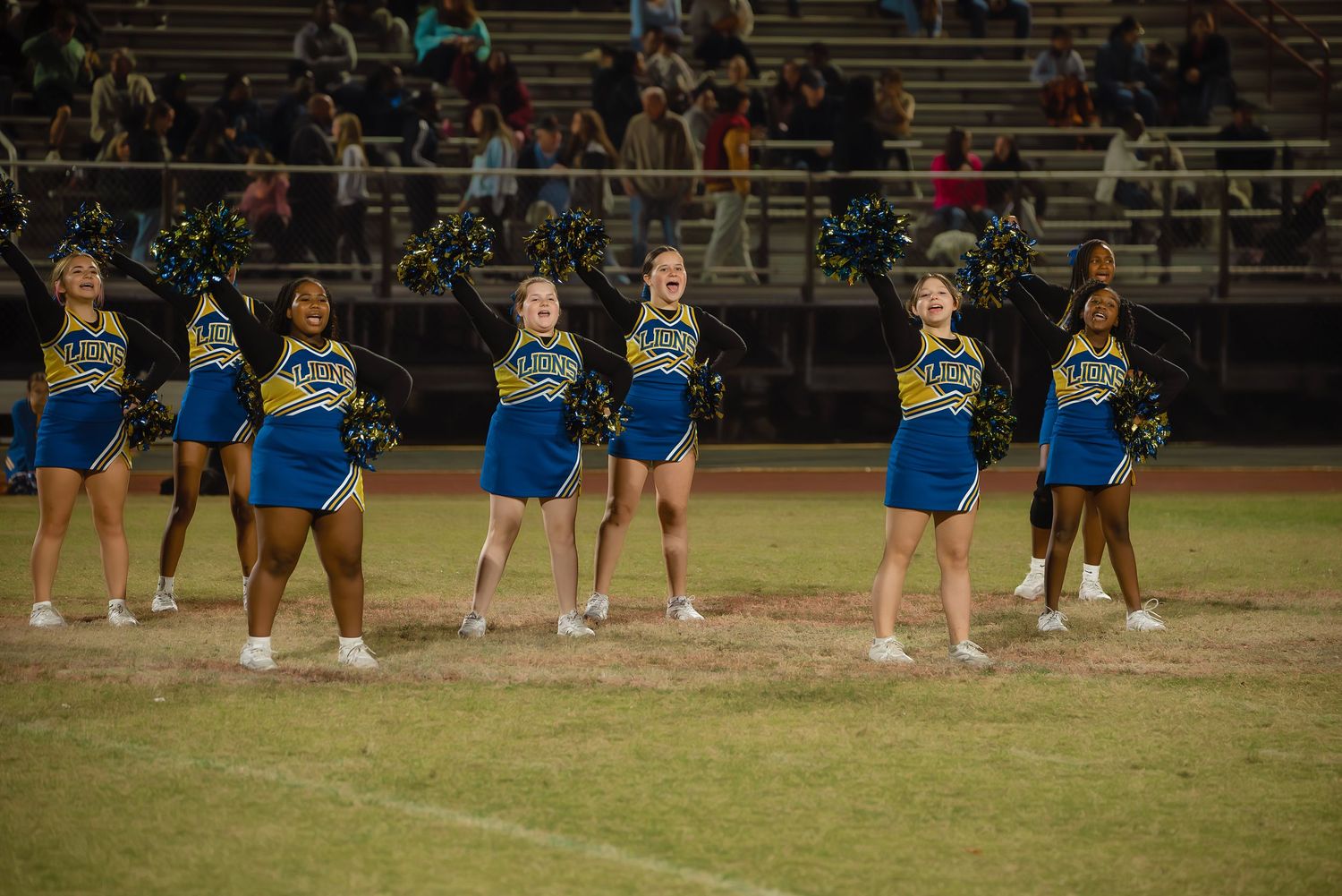 Cheerleaders in blue uniforms perform a routine on a grass field during an evening sports event.