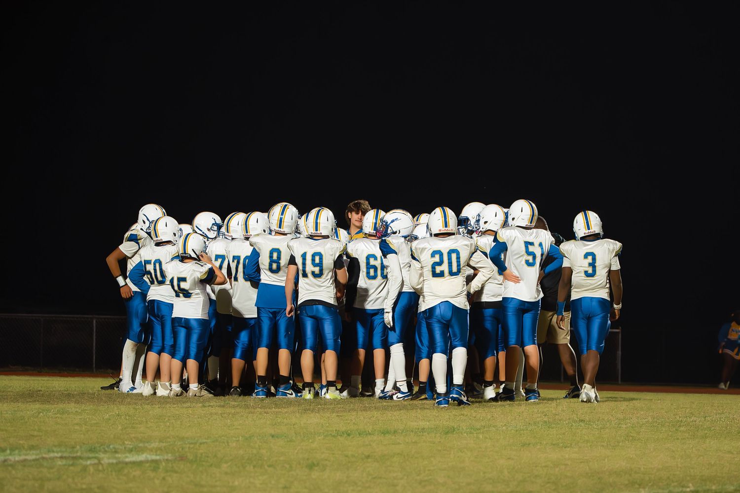Football players huddle together on a dark field during a nighttime game wearing white and blue uniforms.