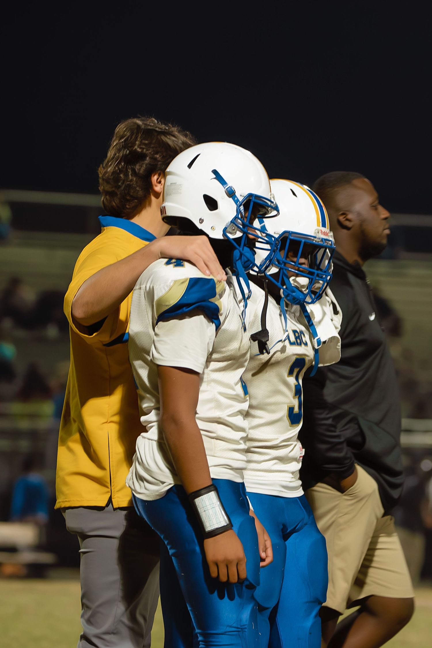Football players in white and blue uniforms stand on sideline with coaches during nighttime game.