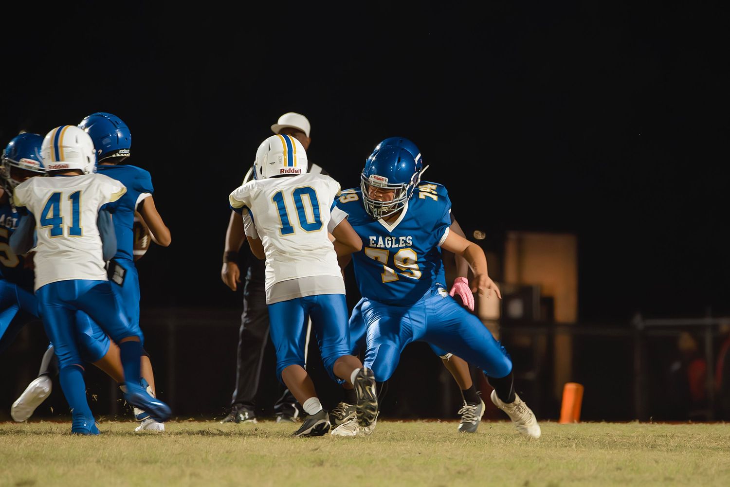 Football players in blue and white uniforms line up in formation during a night game on the field.