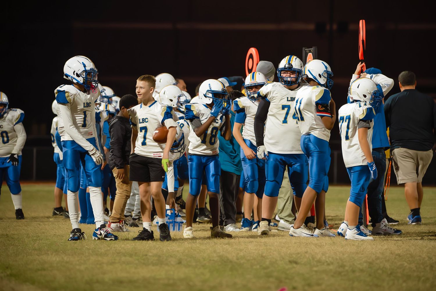 Football players in white and blue uniforms line up along the sideline during a game at sunset.