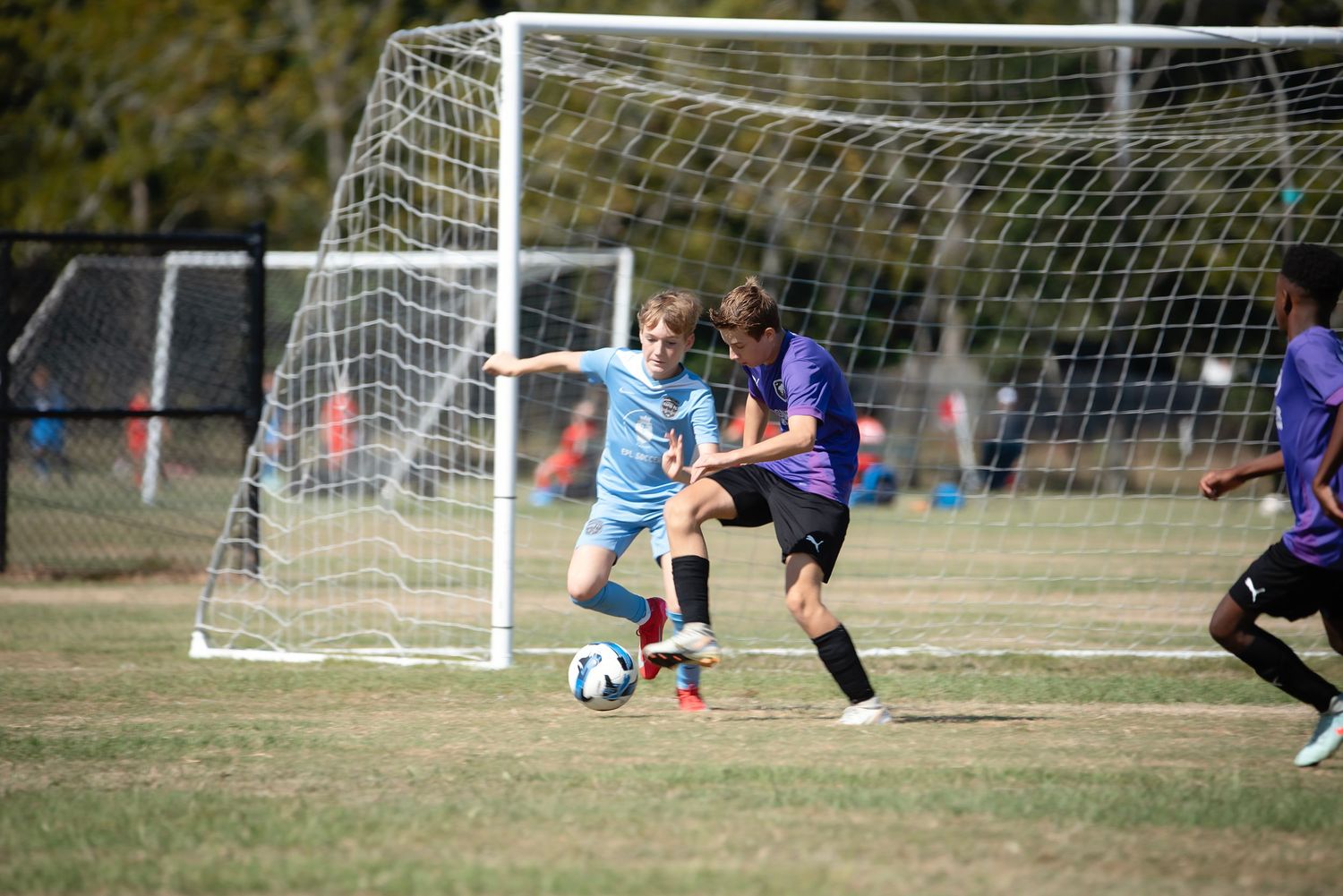 Youth soccer players compete in a match on a sunny day with white goal posts visible in the background.