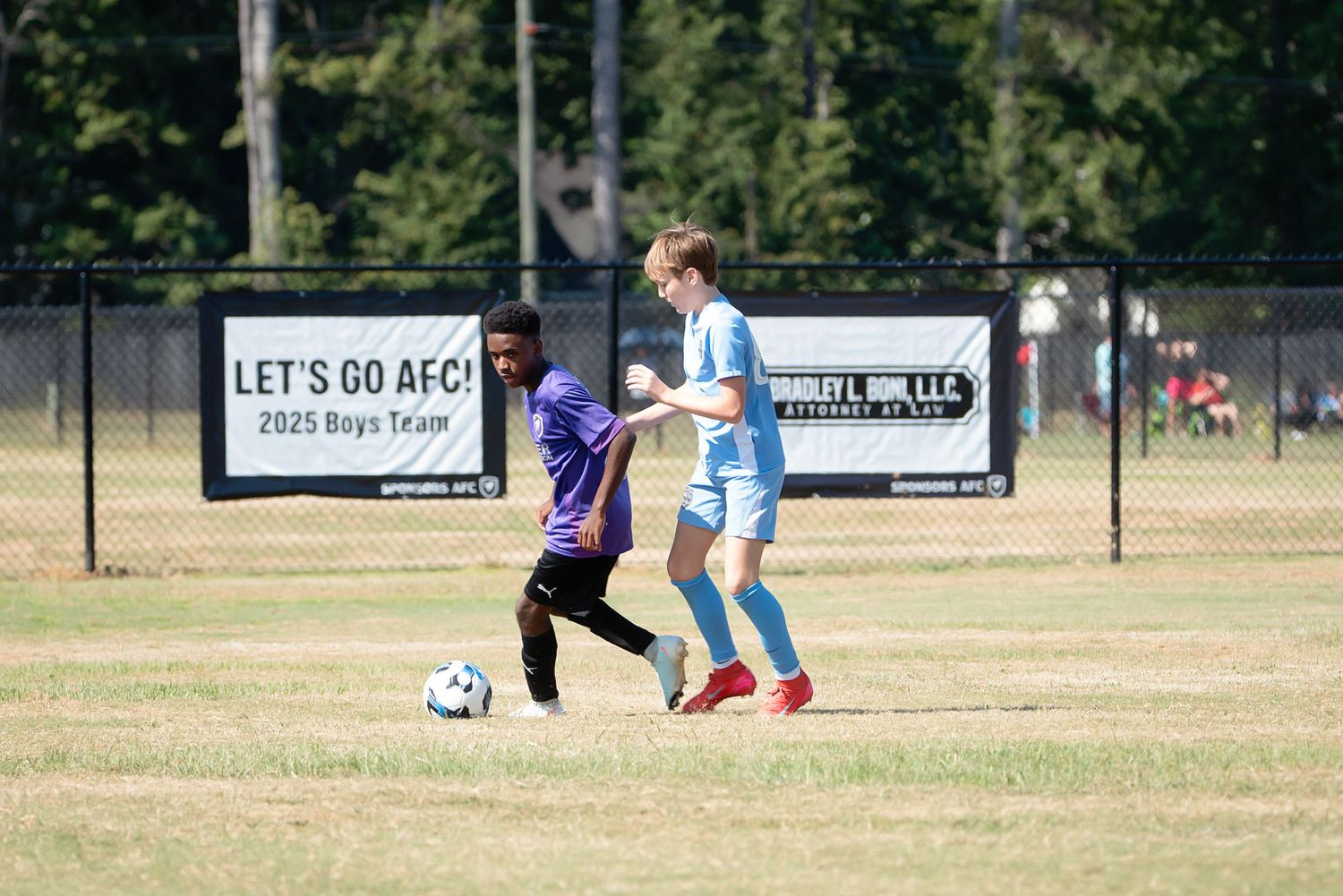 Children play soccer on a dry grass field during a sunny day while others watch from the sidelines.