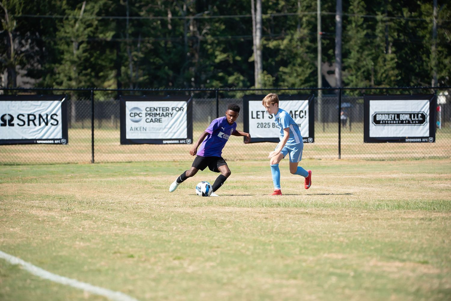 Soccer match in progress with players on field and advertising banners visible along sideline.