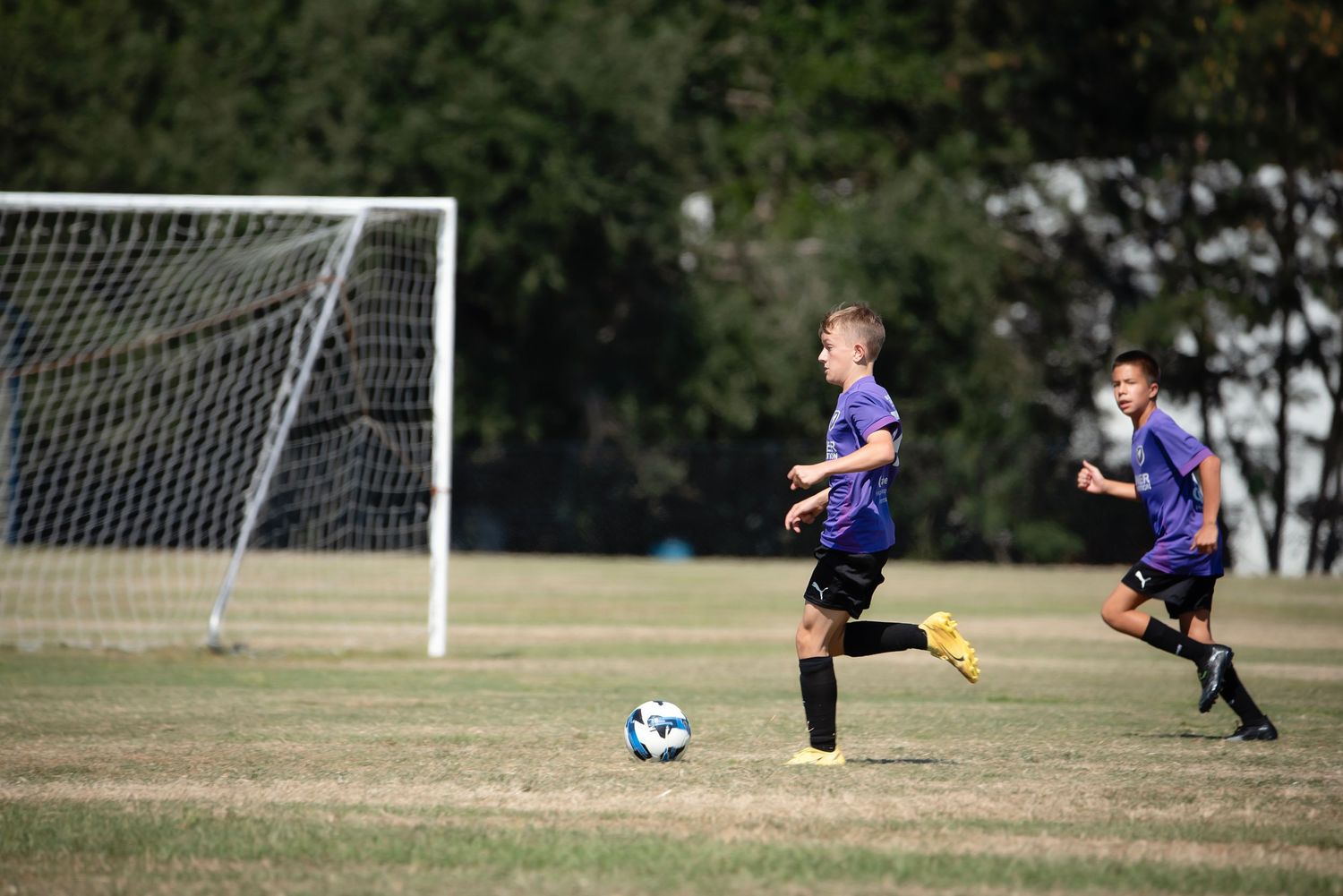 Soccer players in blue and purple uniforms position themselves near a goal on a grass field.