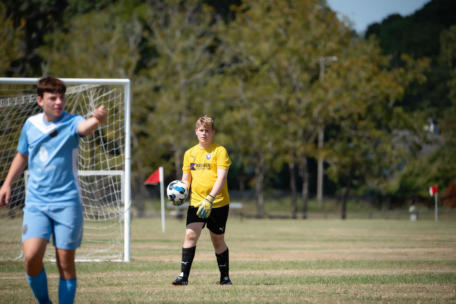 Soccer players in blue and purple jerseys chase after the ball on a sunny field.