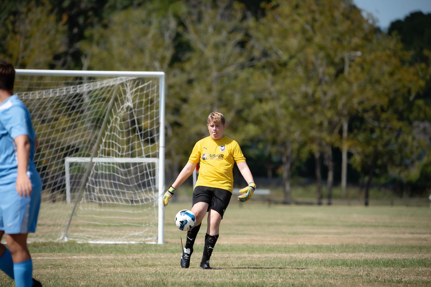 Soccer players compete on a grassy field with goal posts visible in the background during a sunny day.