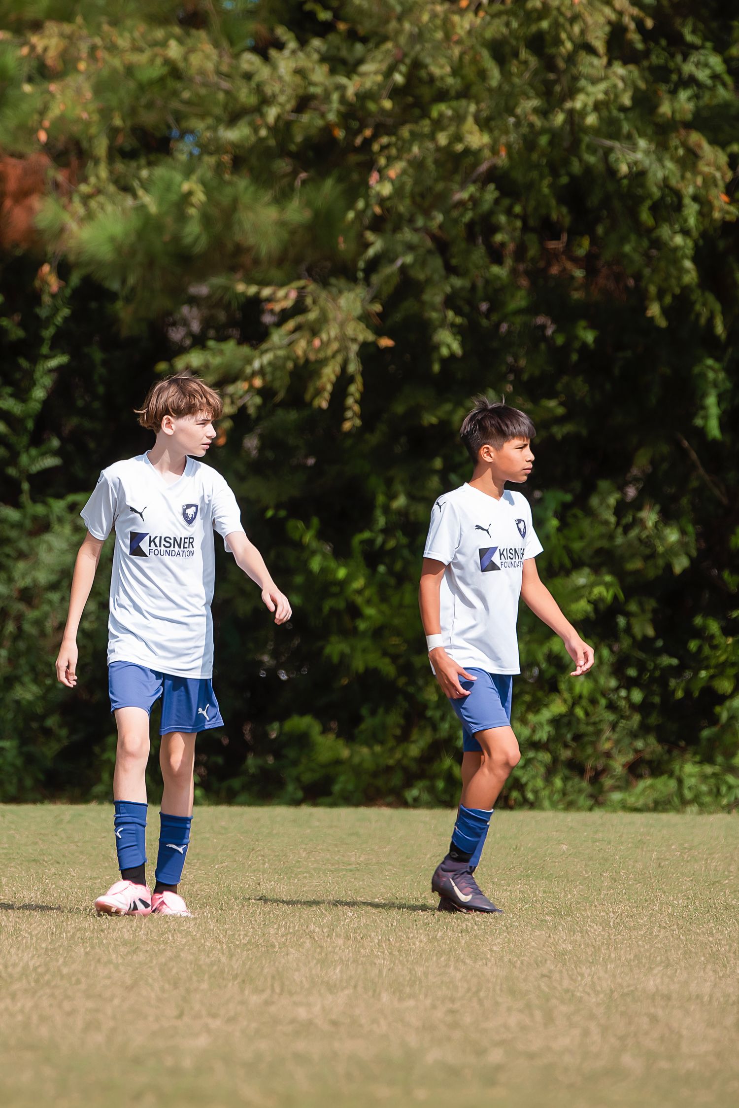 Young soccer players in white uniforms practice drills on a sunny field with trees in background.