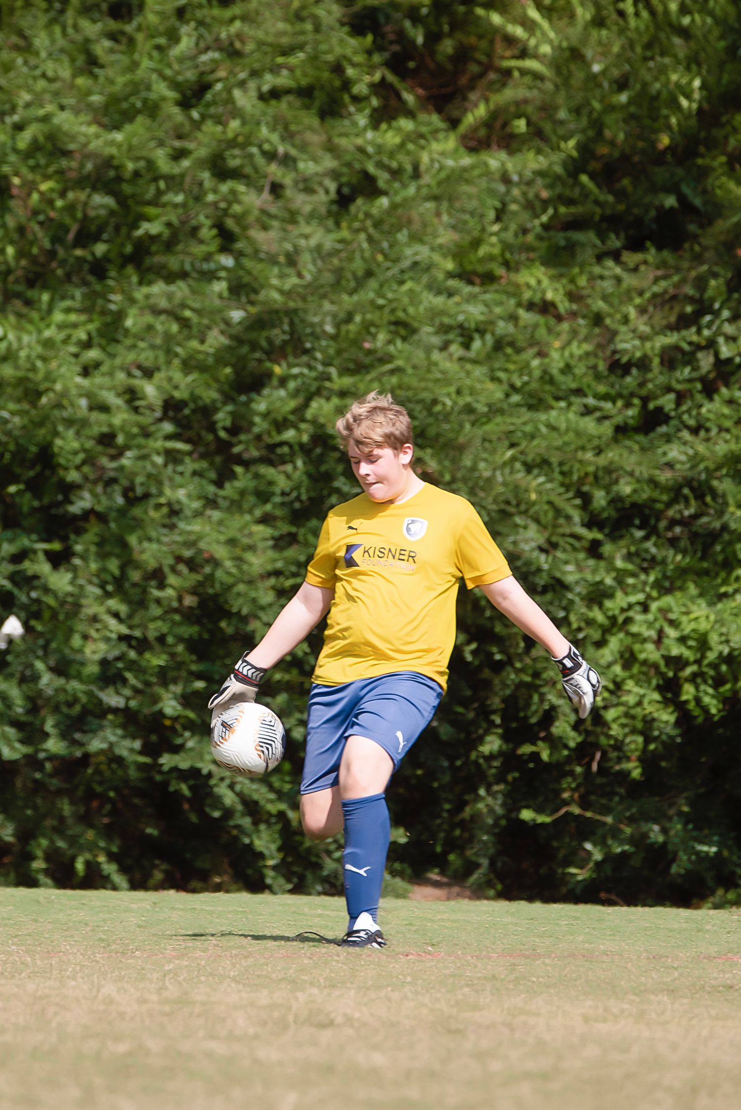 Player in yellow jersey practices soccer ball control drills on grass field near wooded area.
