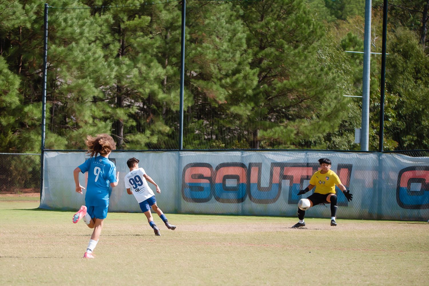 Soccer match action shot showing players competing on a field with trees in the background.