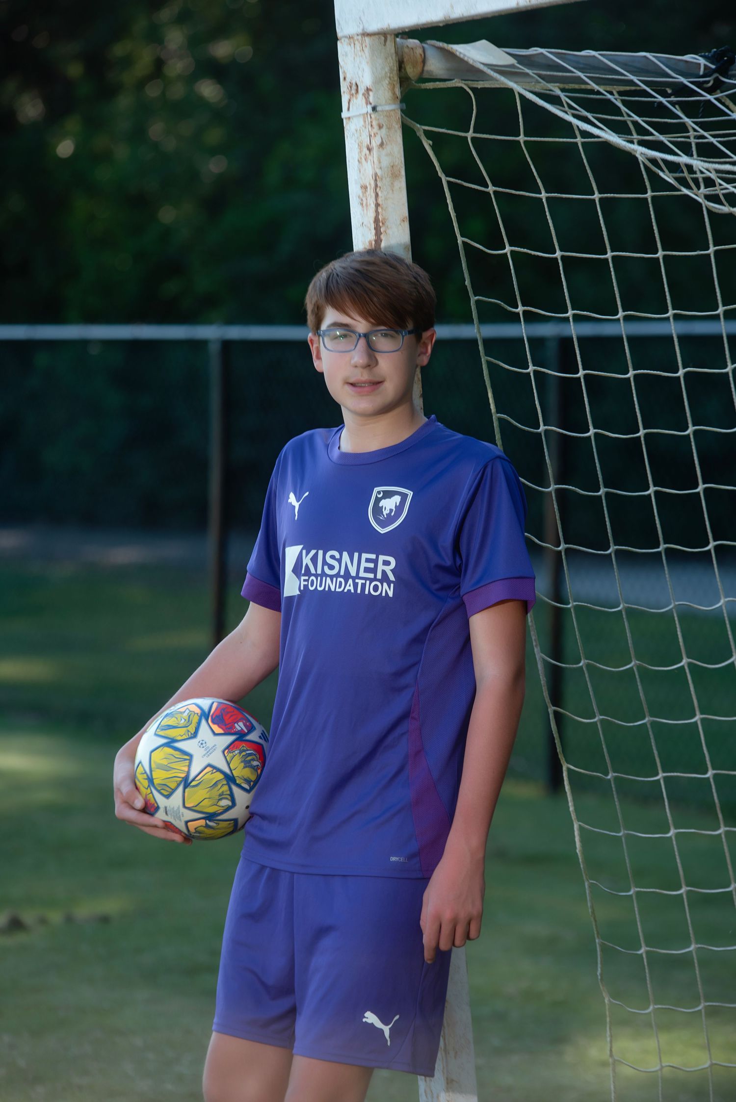 Young soccer players in purple Kisner jerseys pose with colorful soccer balls by a goal net on a grass field.
