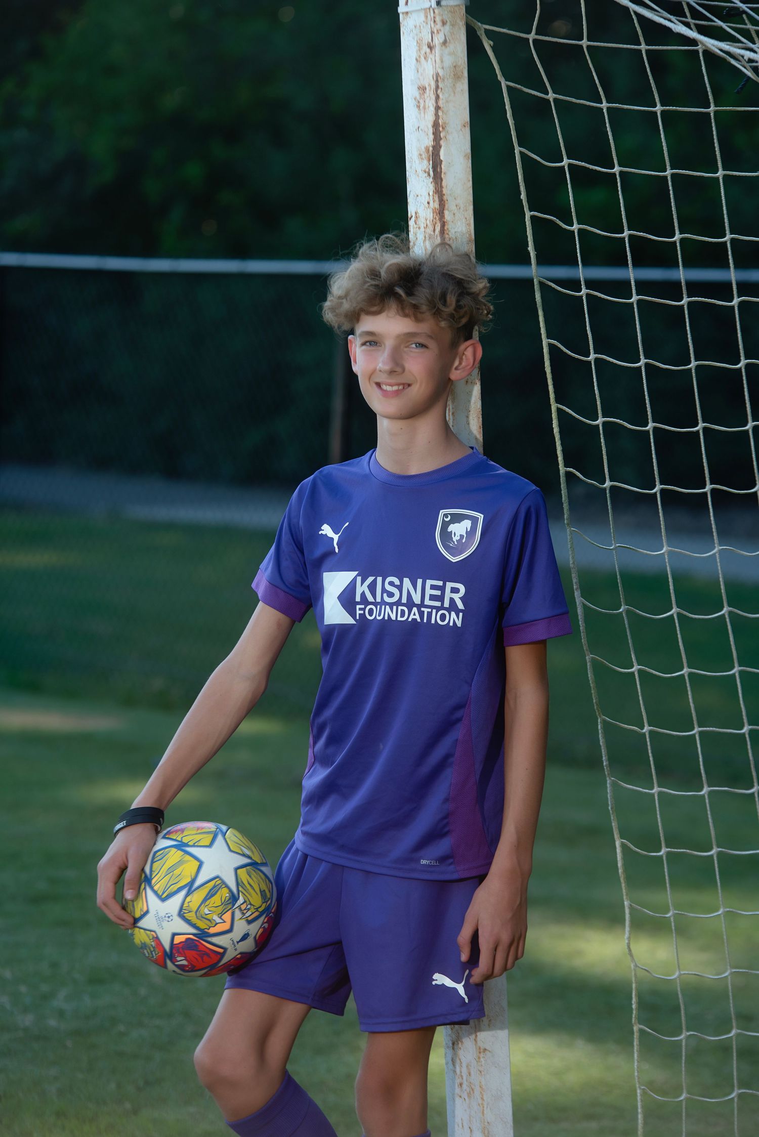 Young soccer player in blue Puma uniform poses with colorful ball next to goal post during outdoor practice session.