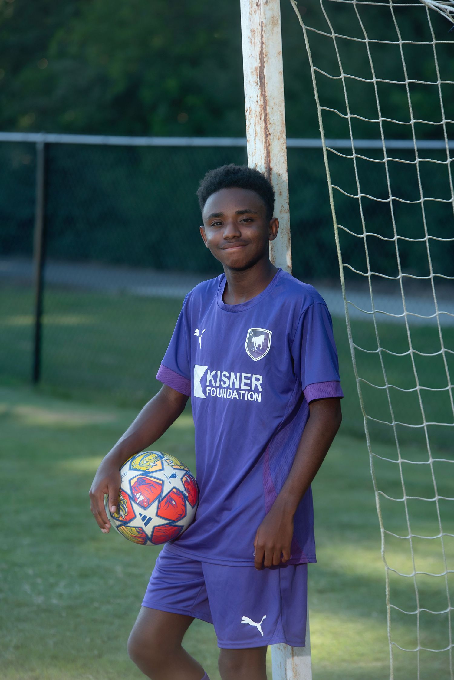 Soccer players in purple uniforms stand near a white goal post holding colorful soccer balls.