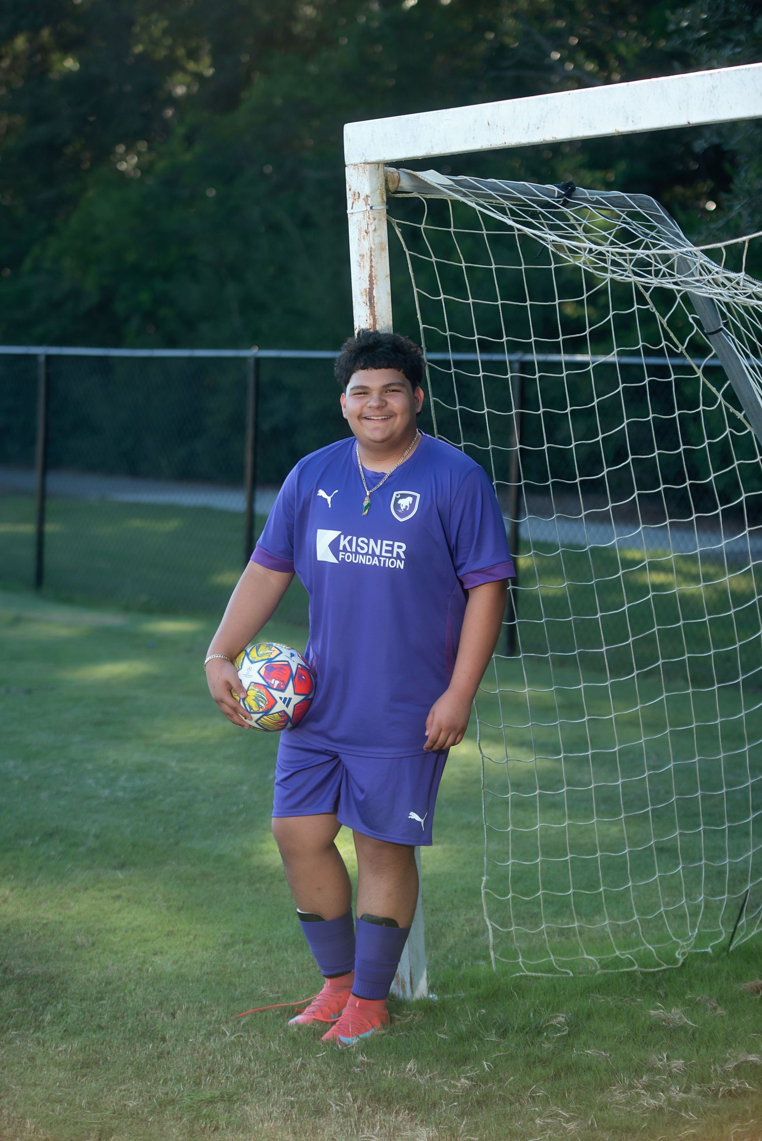 Youth athletes in purple Puma jerseys show off their soccer balls by the goalpost during practice.