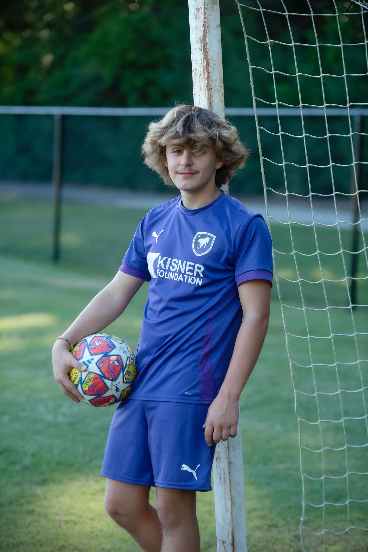 Young soccer player in blue uniform stands near goal post holding a red soccer ball on green grass field.