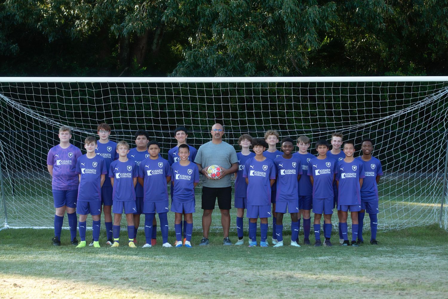 Soccer team in matching purple jerseys poses together in front of a goal net on a grassy field.