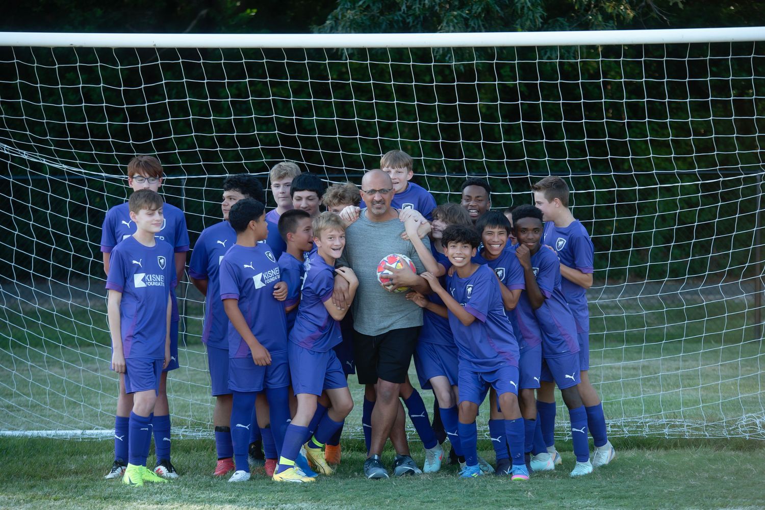 Soccer team in blue jerseys gathers together in front of a goal net on a sunny day.