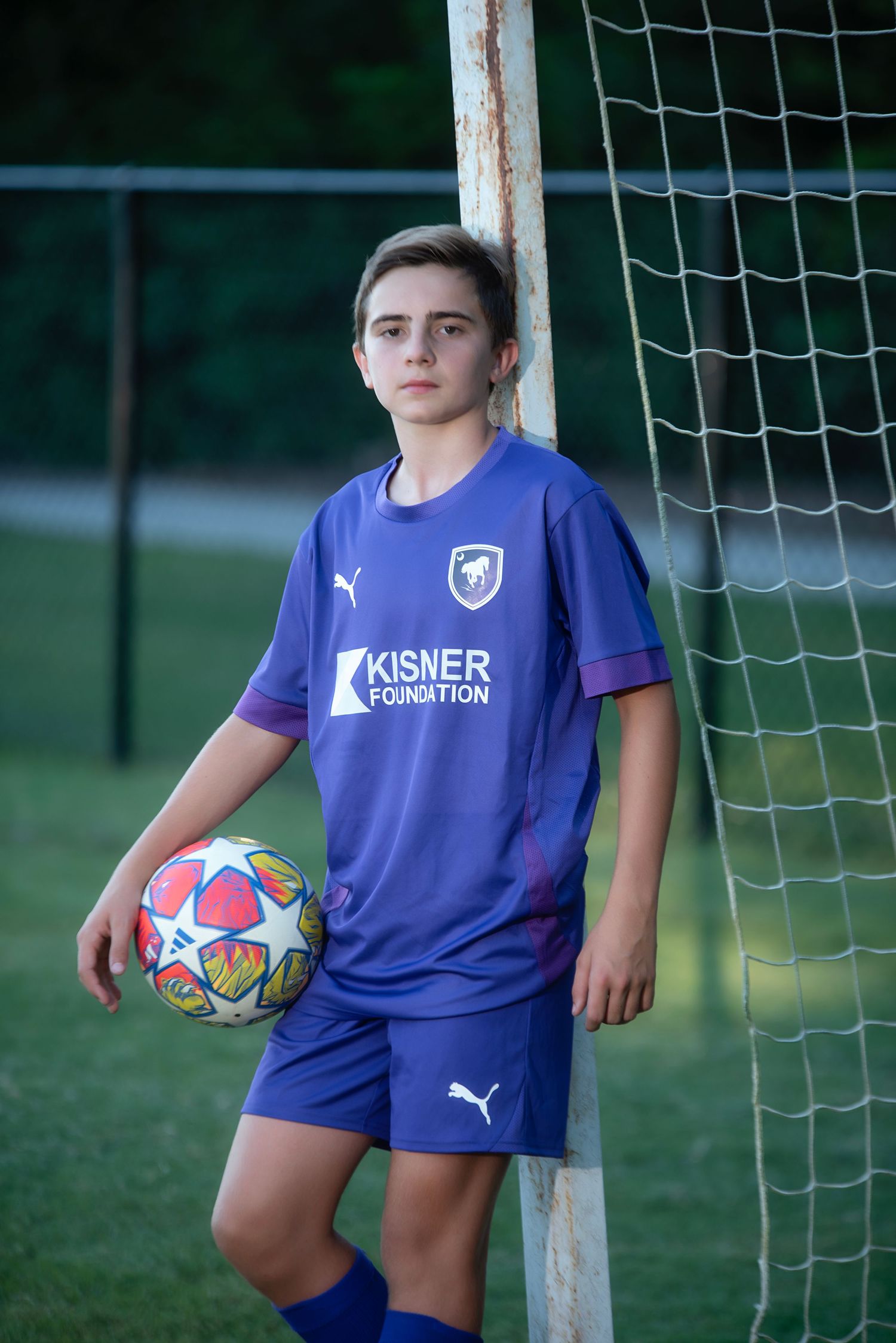 Youth athlete in blue Kisner Foundation uniform holding soccer ball while standing by soccer goal net.