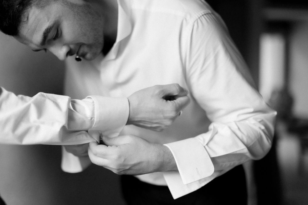 A person in white dress shirt buttoning up formal black leather shoes against a neutral background.
