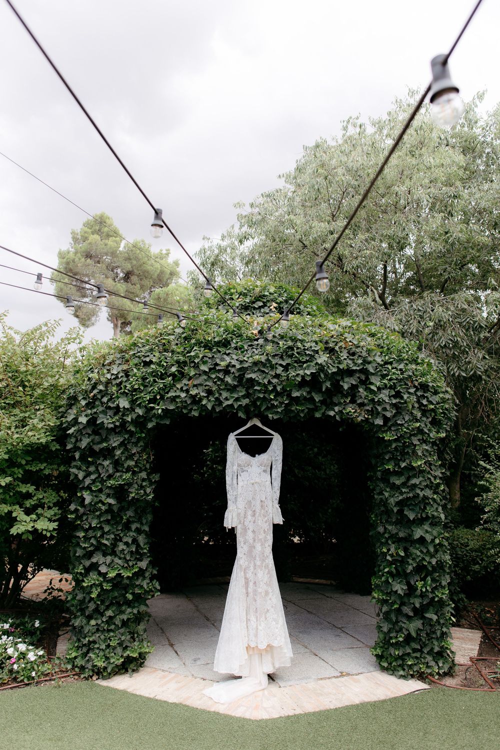 A beautiful white wedding dress hangs under a lush green ivy-covered archway in an outdoor garden setting.