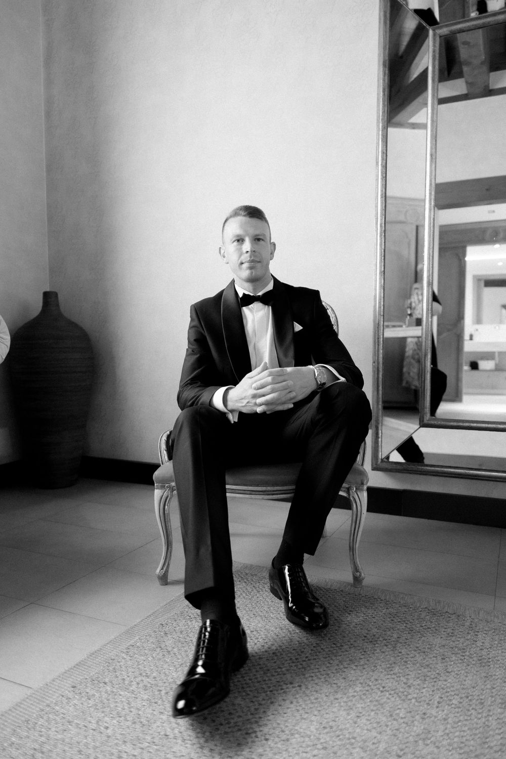 A person in a classic black tuxedo and bowtie sits formally on an elegant chair in a minimalist room.