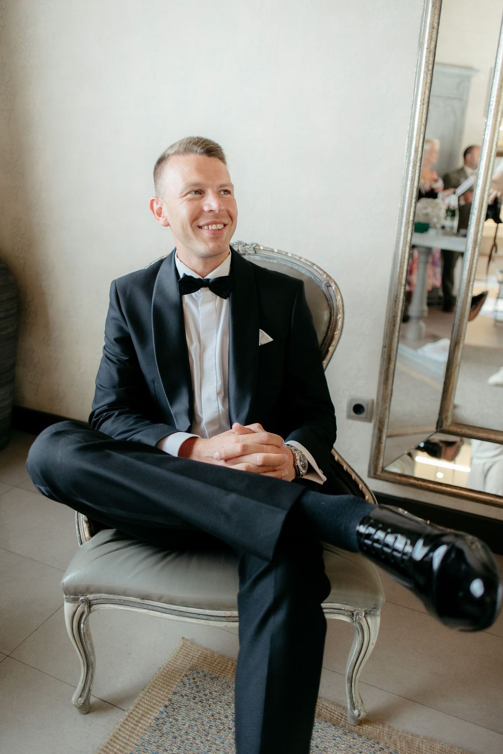 Person in formal black tuxedo sits in elegant vintage chair for wedding preparation portraits.