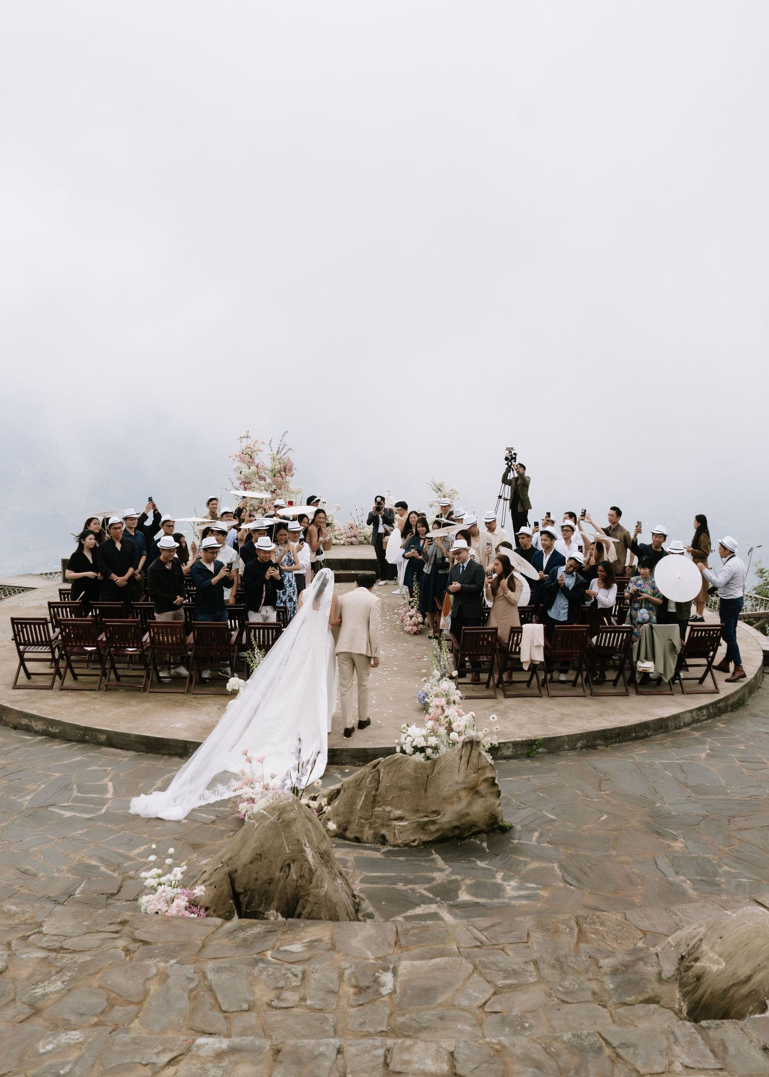 Aerial view of a bride in a long veil walking toward the altar at a misty outdoor wedding ceremony with seated guests.