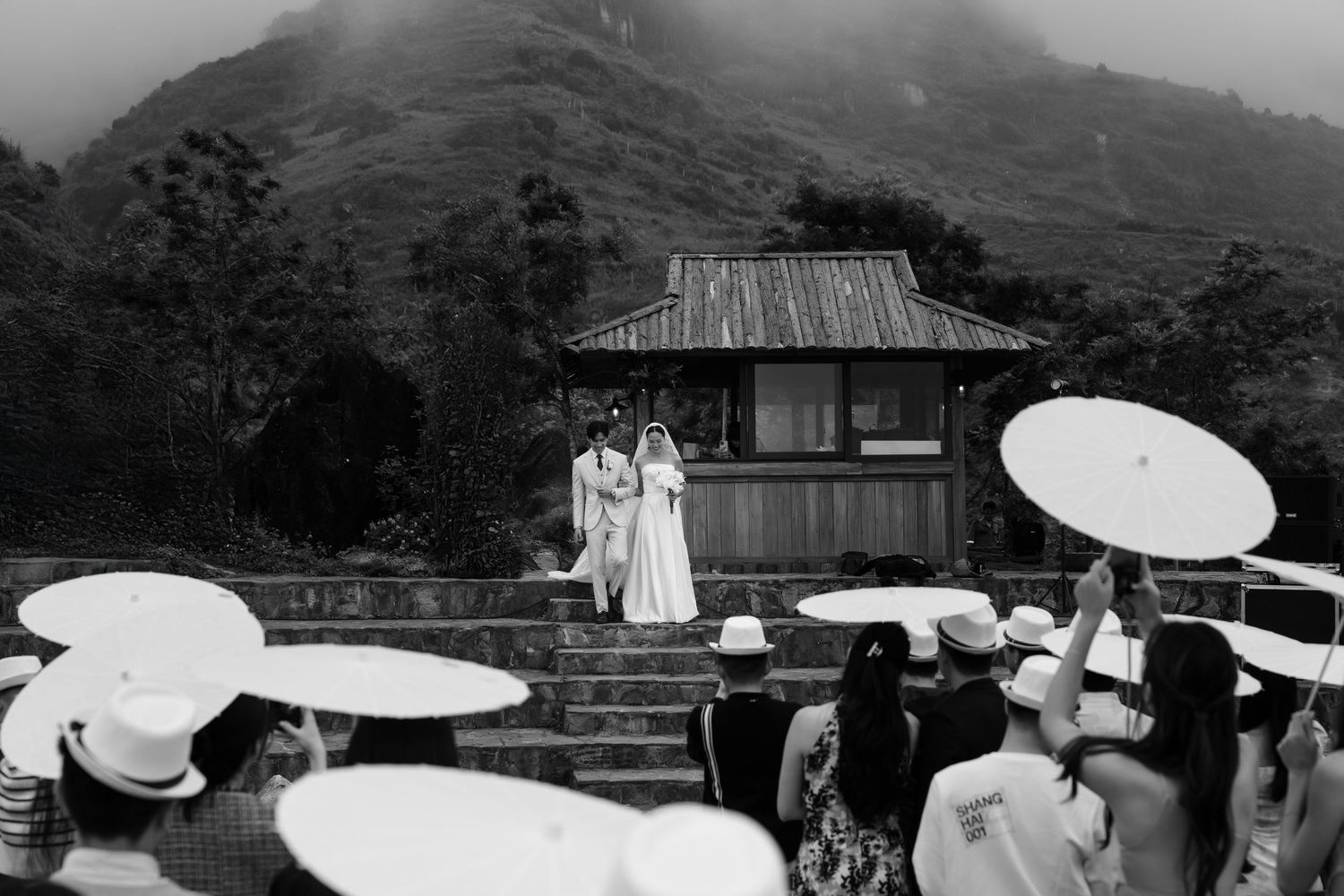 Black and white photo of a couple standing on steps before a rustic building as guests hold white parasols below.