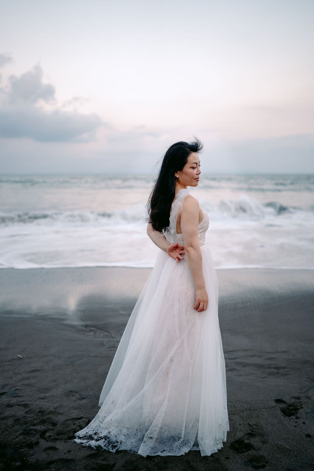 Person in flowing white dress poses on black sand beach with crashing waves.