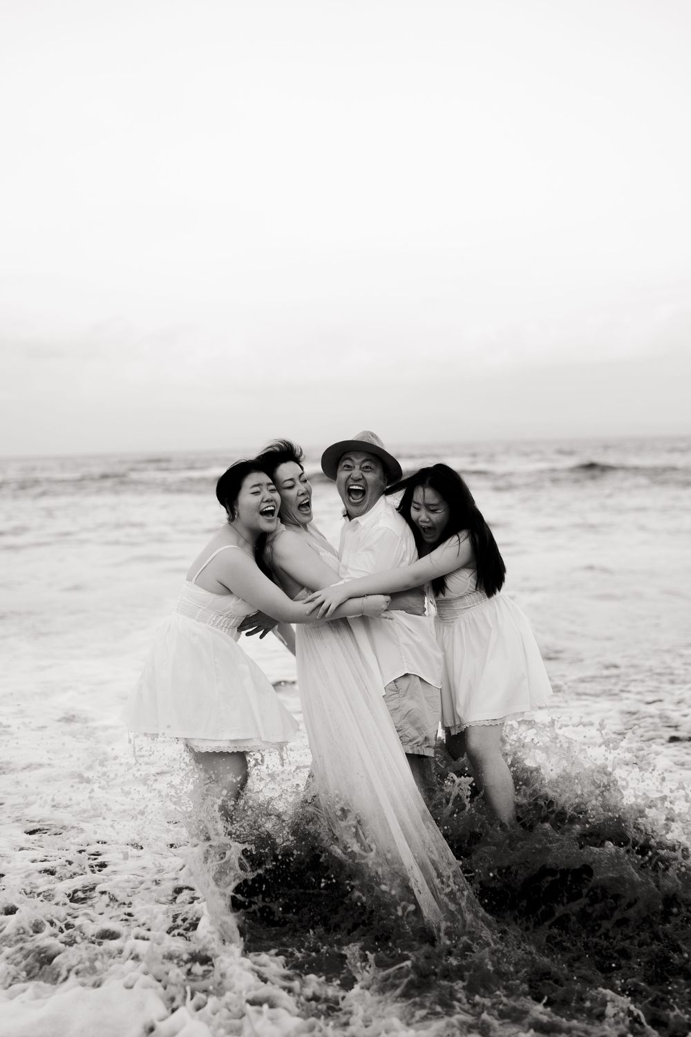 Three people in white dresses laugh together as ocean waves splash around them on the beach.