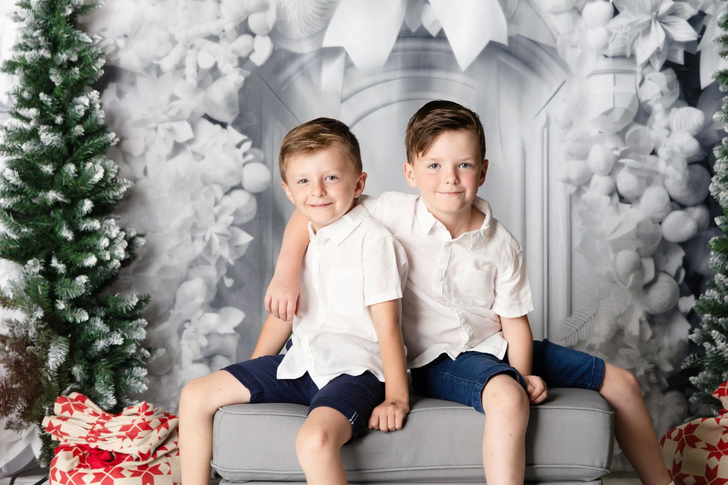 Children sitting together on a couch surrounded by Christmas decorations and a white holiday backdrop.