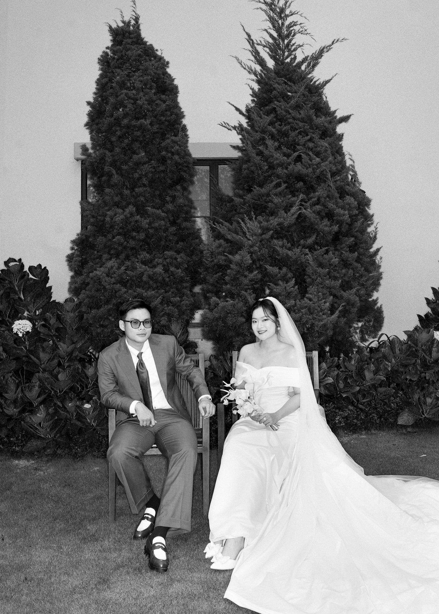 Black and white vintage-style wedding portrait of bride and groom seated outdoors among tall evergreen trees.