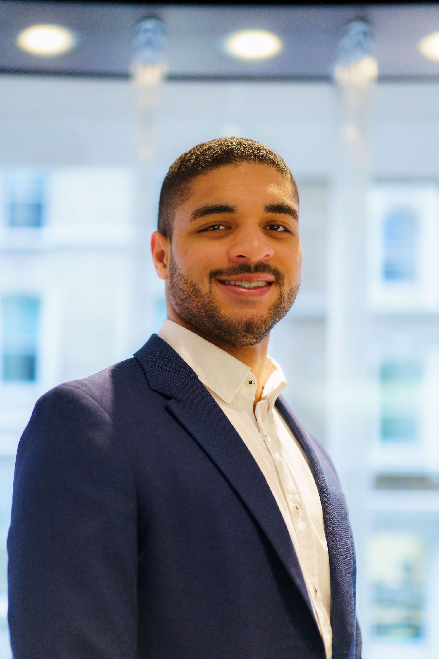 Young man smiling for his headshot indoors, with a glassy building in the background